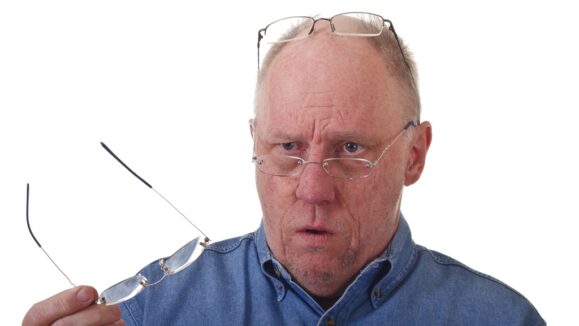 An older balding man in blue denim shirt with three pairs of reading glasses